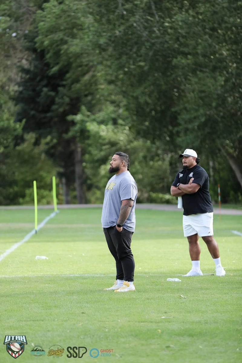Two coaches in casual shirts stand together watching play from the sideline