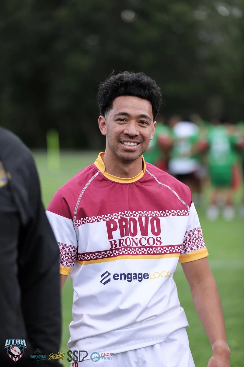 Young Provo Broncos player in a maroon and white jersey smiles before the game with green-clad opponents blurred behind him