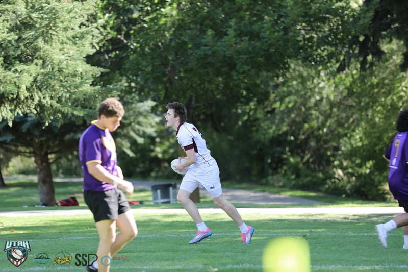 A Riverton Seagulls player in white and maroon runs upfield with the ball past a Lehi Raiders defender in purple.