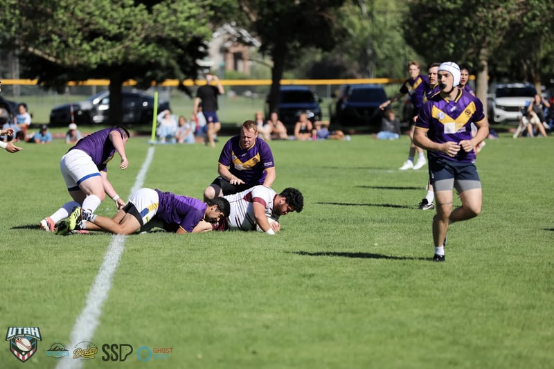 A scrum-capped Lehi Raiders player runs off a ruck as both teams contest the ball on the ground.