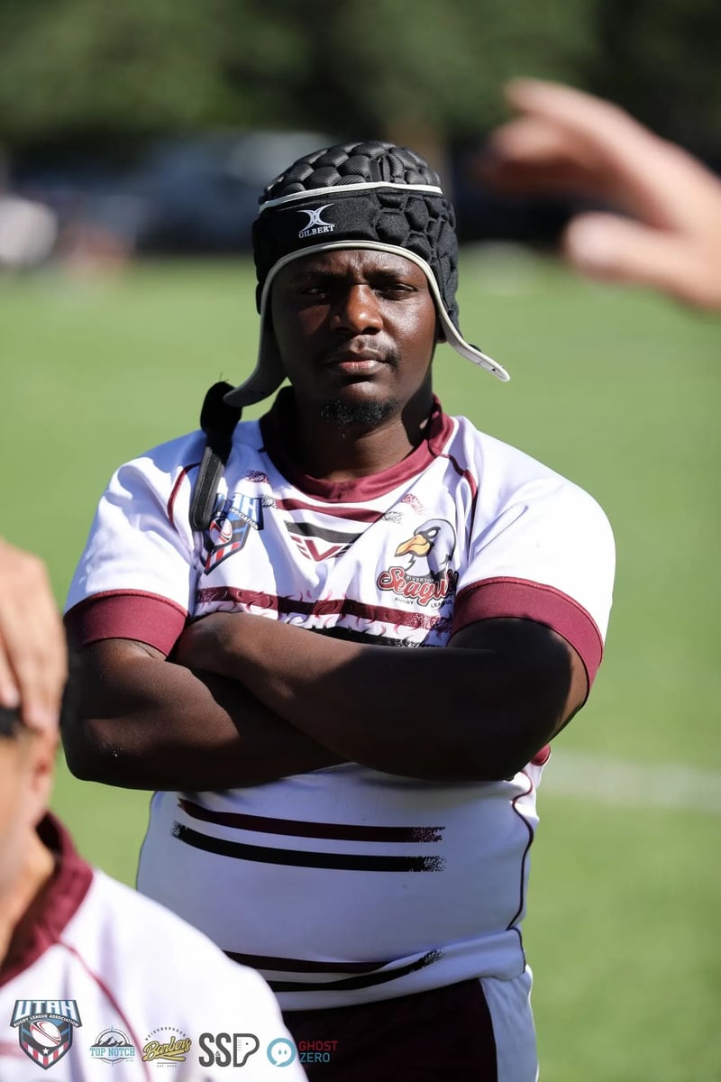 Seagulls player in white and maroon stands with arms folded and headgear strapped on, listening at a sideline huddle.