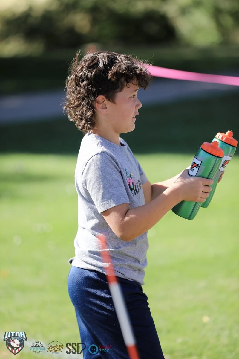 Young ball boy in a gray t-shirt carries an armful of water bottles along the sideline of the rugby pitch.