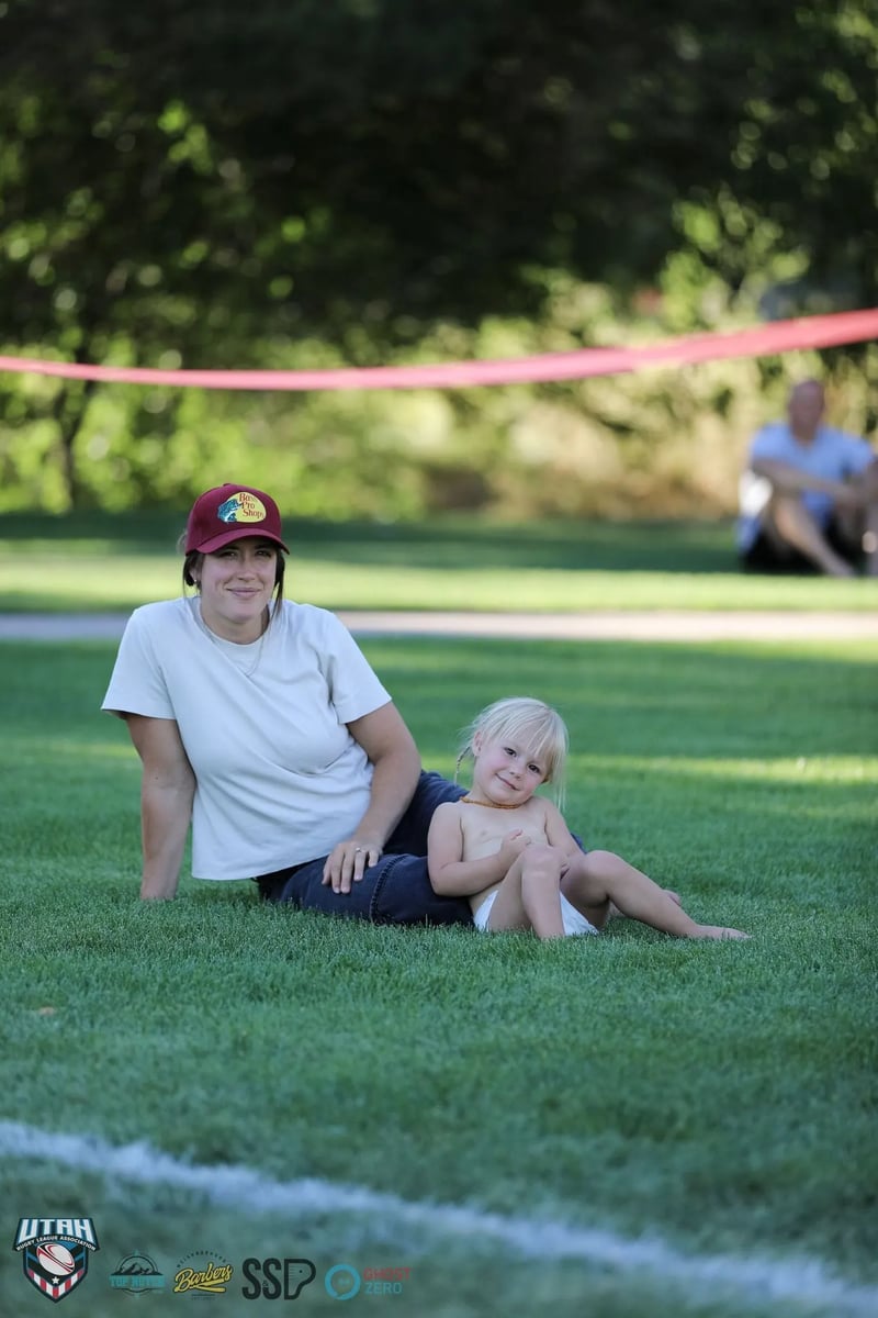 A woman in a maroon cap sits on the sideline grass with a smiling toddler, watching the match from the shade.