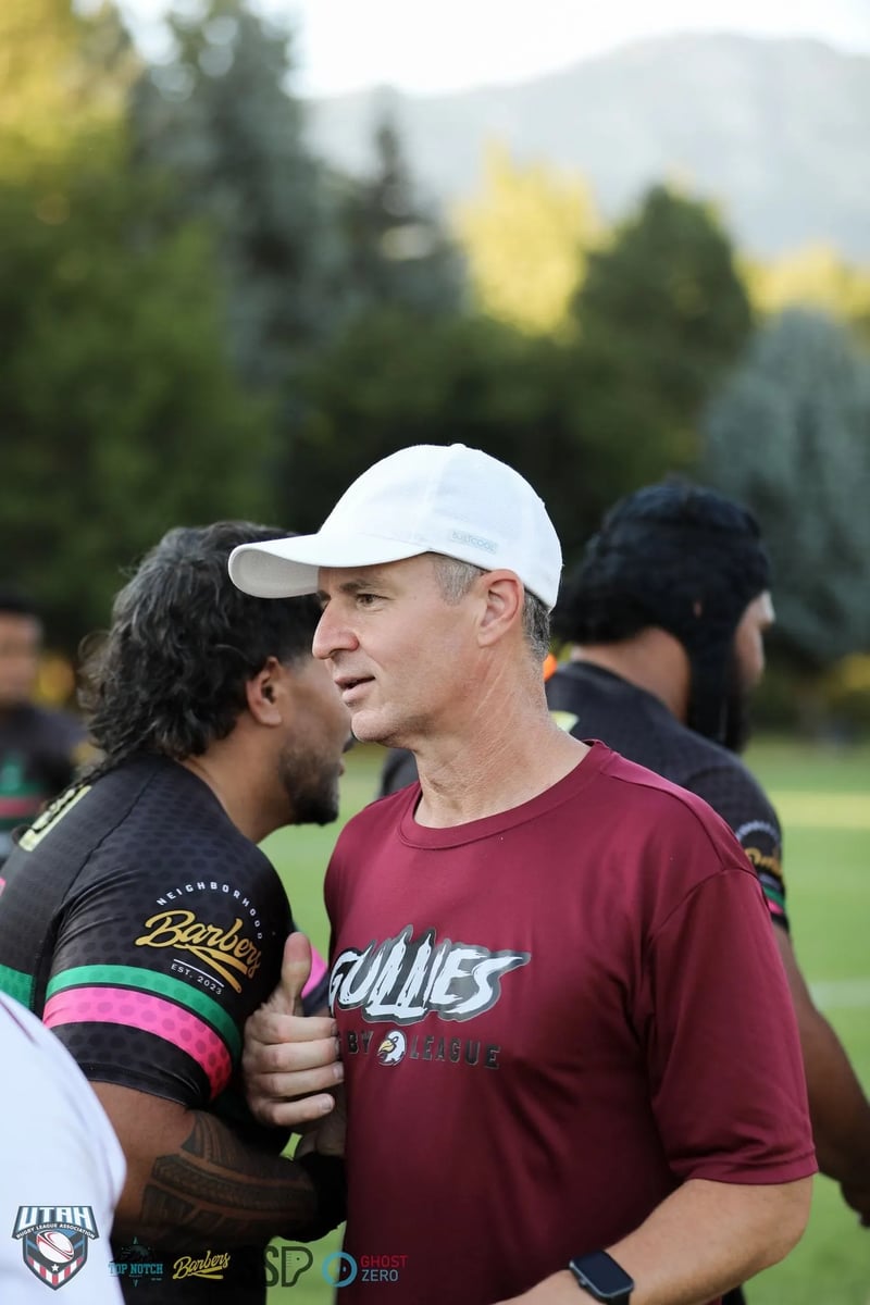 A coach in a white cap talks face to face with a dark-kitted player amid the post-match handshakes in warm evening light.
