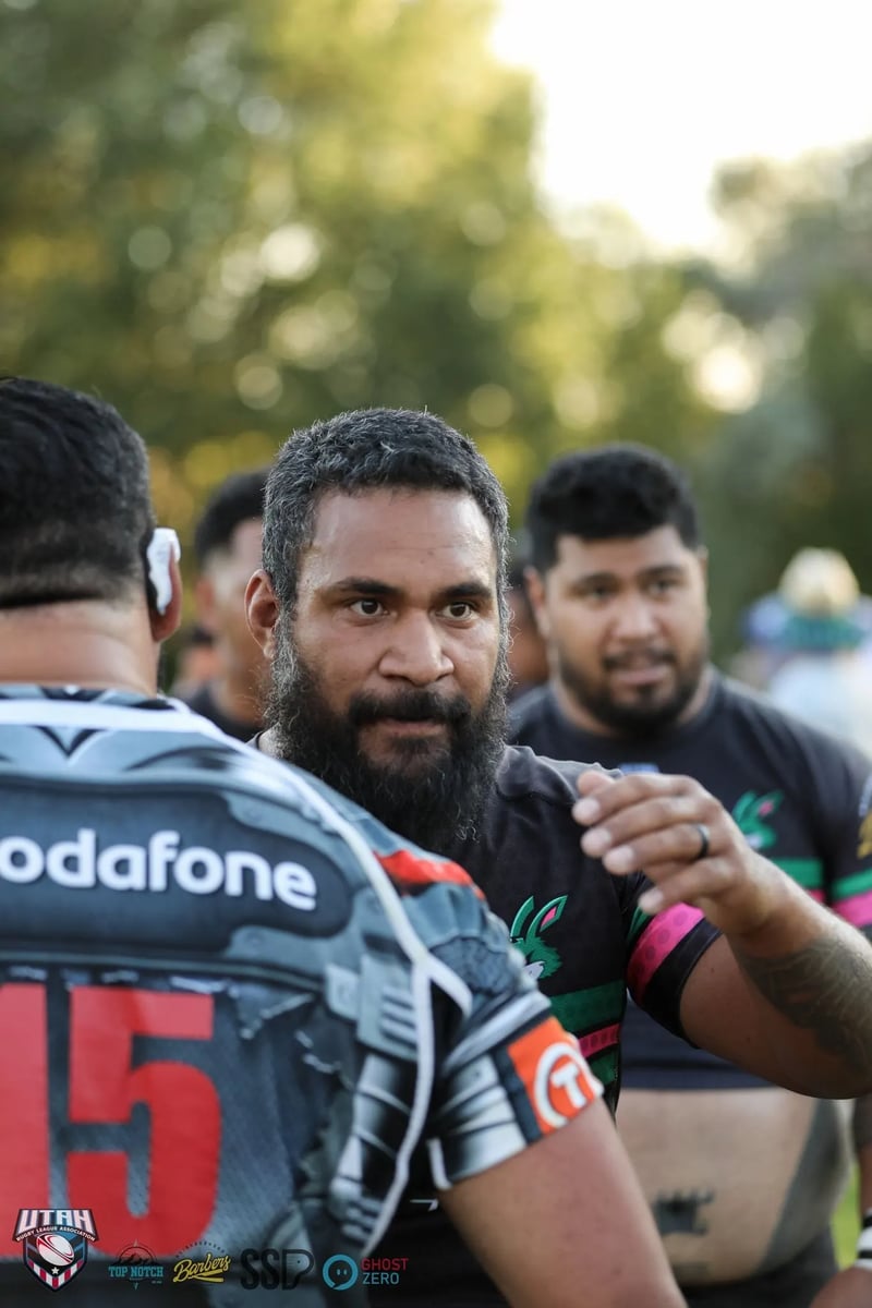 A bearded man in a Vodafone Warriors jersey grins intensely amid the post-match mingling on the field.