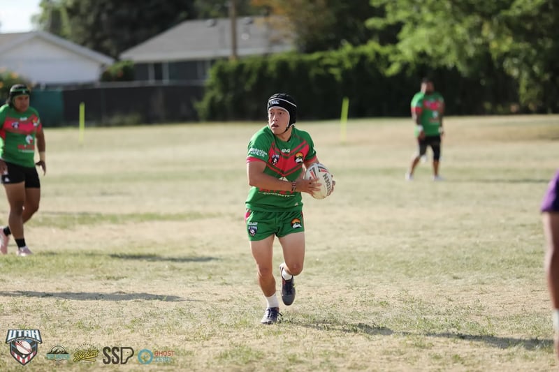 Rabbitahz player in green and red wearing a black scrum cap carries the ball forward in open space, teammates spread across the dry field around him.