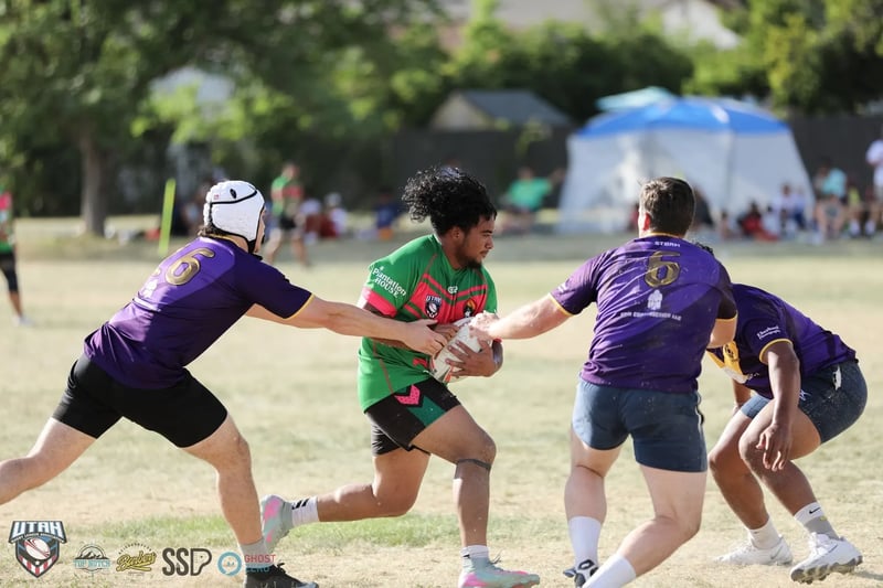 Rabbitahz ball carrier in green and red splits two stretching Raiders defenders in purple, one in a white scrum cap, with a festival tent and sideline spectators in the background.