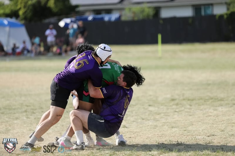 Two Raiders defenders in purple, one wearing a white scrum cap, wrap a Rabbitahz ball carrier in green in a heavy double tackle, festival spectators visible along the far sideline.