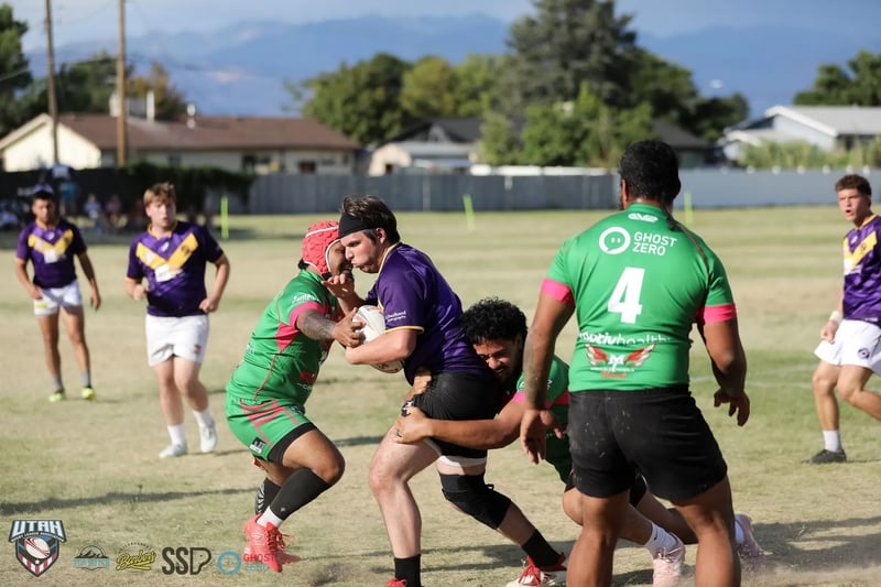 Two Rabbitahz defenders in green and red halt a Raiders ball carrier in purple mid-stride, a teammate wearing number 4 backing up the tackle with mountains on the horizon.