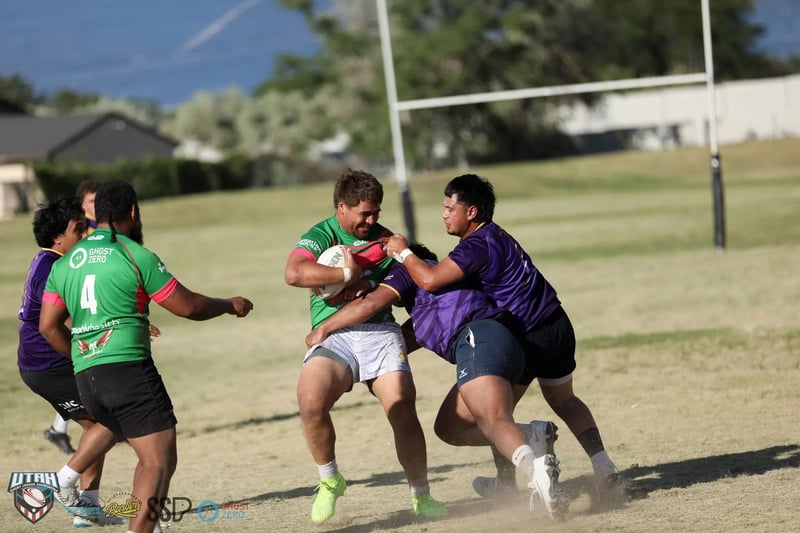 Rabbitahz forward in green and red drives with the ball into a Raiders tackler in purple, dust kicking up under their feet with the goalposts and hillside behind.