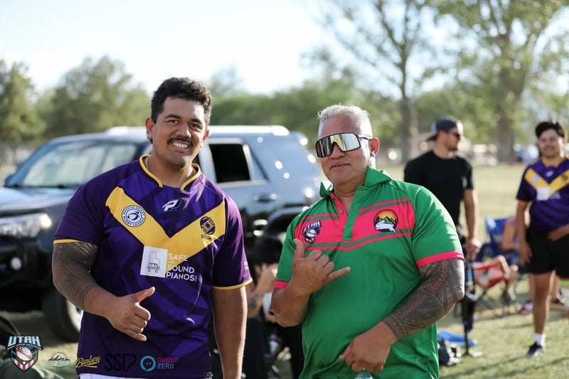 A Raiders player in purple and gold and a player in a green festival jersey pose together throwing shakas beside parked cars at the sideline