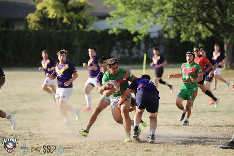 A Rabbitahz ball-carrier in green and red bursts between two Raiders defenders in purple, dust kicking up off the dry ground.