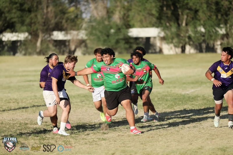 Two Raiders defenders in purple and gold combine to wrap up a barrel-chested Rabbitahz ball-carrier in green in low evening sun.