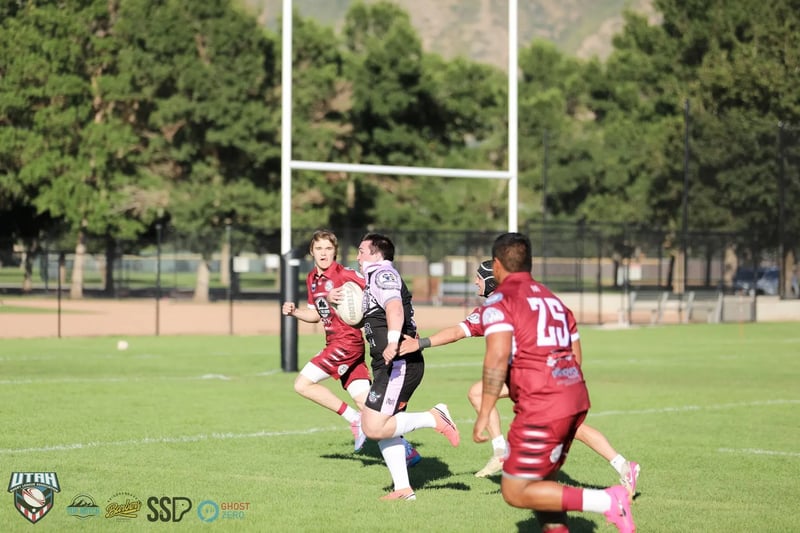 A Lehi Raiders ball carrier in black and lavender attacks between two Riverton Seagulls defenders in maroon, with the goalposts and the foothills behind.