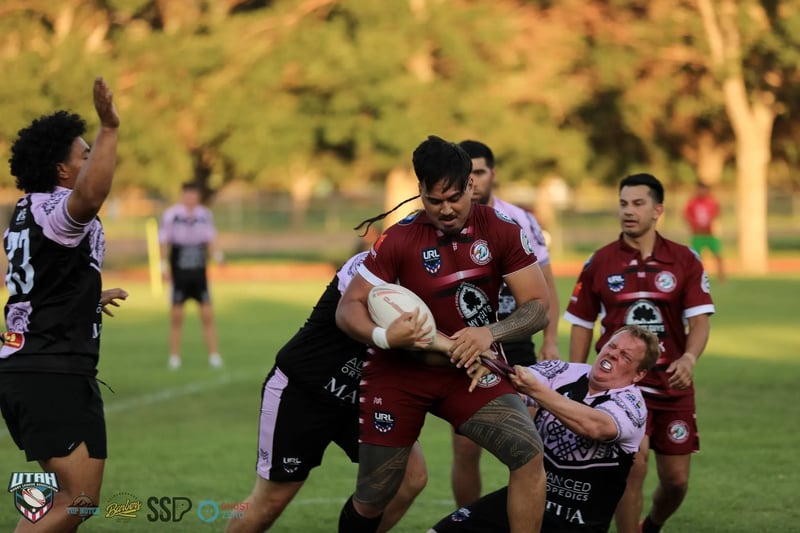 A Riverton Seagulls forward in maroon drives forward with the ball in both hands while two Lehi Raiders defenders hang off him.
