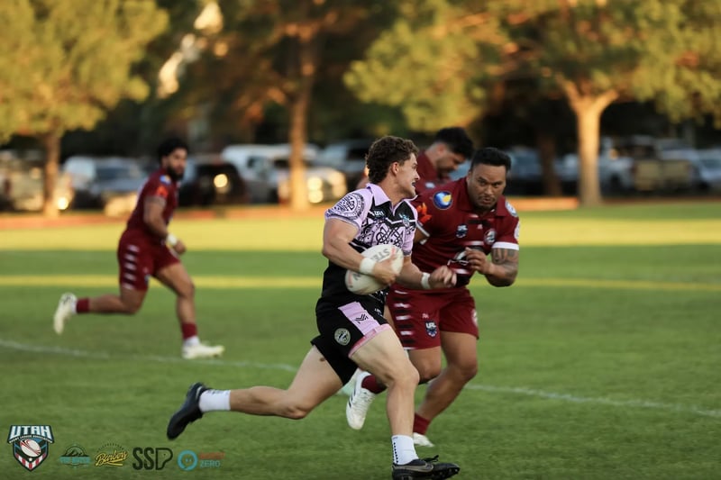 A Lehi Raiders ball carrier runs at the camera with the ball in two hands, breaking clear of a Riverton Seagulls defender reaching for his back.