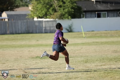 Raiders player in purple sprints away on a breakaway with ball in hand, festival tents and crowds lining the far touchline