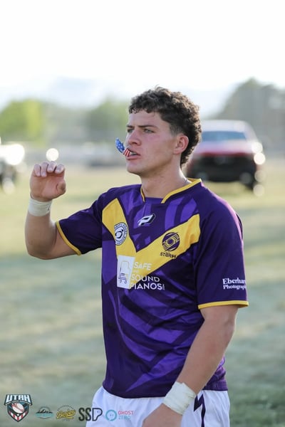 Portrait of a Raiders player in the purple and gold V jersey lifting his mouthguard to his mouth, teammates blurred behind him in warm evening light.