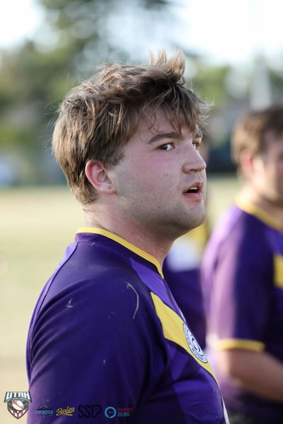Tight golden-hour portrait of a Lehi Raiders player in a purple and gold jersey glancing back over his shoulder between plays.