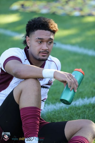 An exhausted Seagulls player in white and maroon sits on the grass with a water bottle in hand and taped wrist, catching his breath after the match.