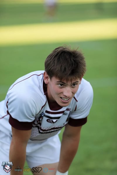 Tight portrait of a young Riverton Seagulls player in a white jersey with maroon trim, bent forward with an intense expression during a break in play.